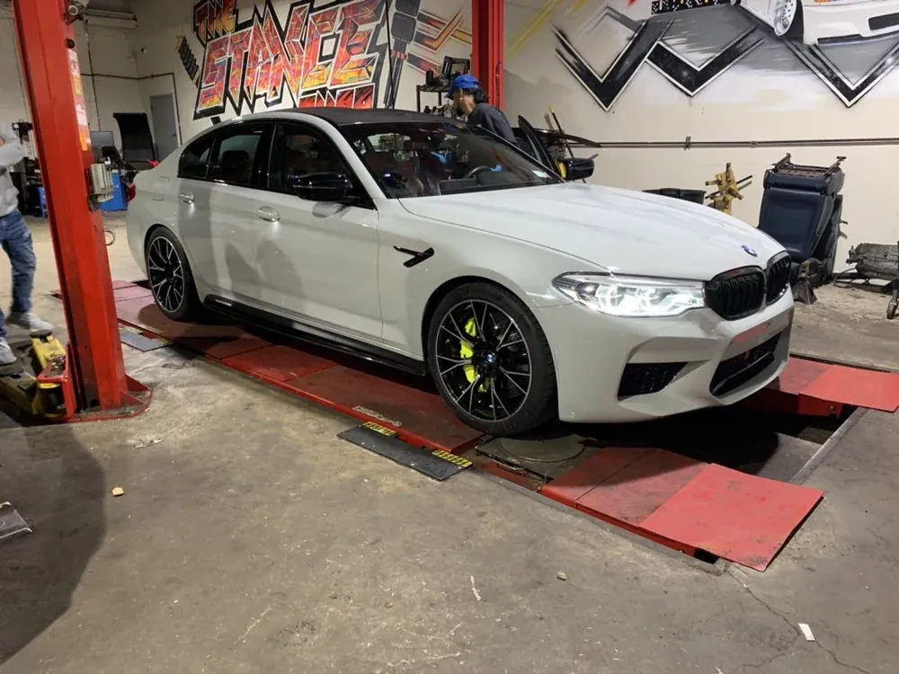 A white BMW M5 on the alignment rack beside The Stance Shop mural