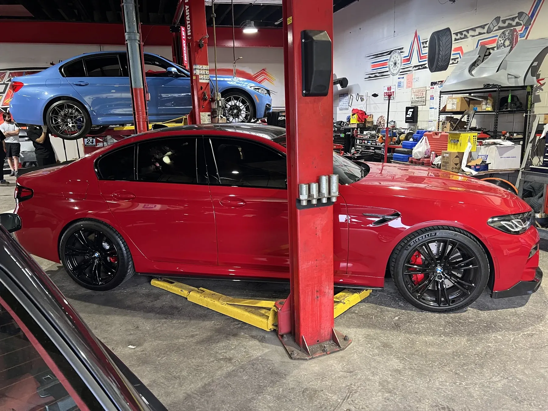 A red BMW M5 inside The Stance Shop with a blue BMW M3 on the lift behind it